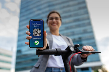 Young businesswoman on the electrical scooter is showing digital immunity passport on the mobile phone.