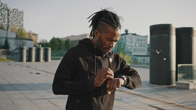 Active african american guy jogging in urban area, turning on activity tracker on smartwatch and start running