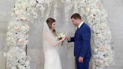 beautiful and happy bride and groom exchange rings in an arch of flowers