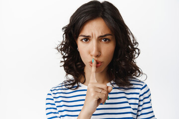 Fototapeta premium Close up of woman shushing with angry face, scolding tell to keep quiet, hush gesture, frowning with disapproval, standing in t-shirt against white background