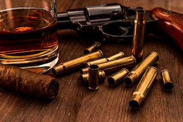 Empty shells from the weapons with glass of whiskey and revolver with cuban cigar on a wooden table