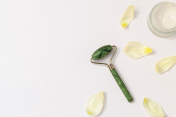 Jade face roller for facial massage with petals on white background, top view with copy space