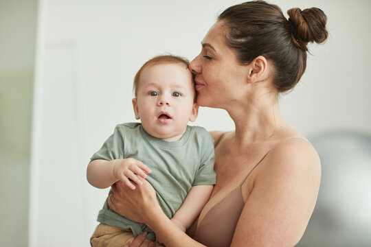 Candid portrait of loving mother kissing baby boy looking at camera in cozy home interior, copy space