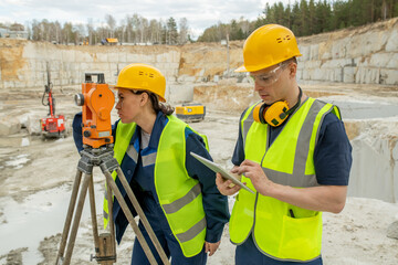 Female engineer using geodetic station while her colleague scrolling in tablet