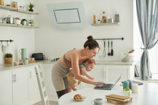 Portrait of modern mother with disability holding baby while using laptop in kitchen interior, copy space