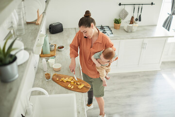 High angle portrait of modern mother with disability holing baby while cooking in cozy kitchen...