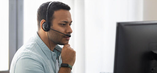 Focused young Indian man with in headphones working at laptop at home office, call center employee,...