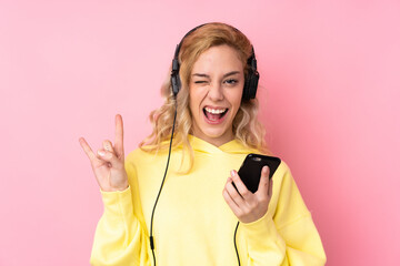 Young blonde woman wearing a sweatshirt isolated on pink background listening music with a mobile making rock gesture