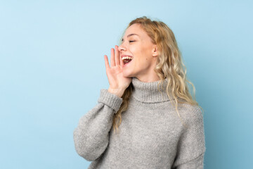 Young blonde woman wearing a sweater isolated on blue background shouting with mouth wide open to the lateral