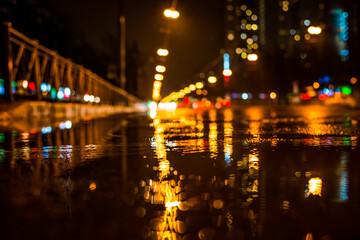 Fototapeta na wymiar Rainy night in the big city, approaching headlights of cars traveling along the avenue. View from puddles on the pavement level