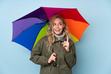 Young blonde woman holding an umbrella isolated on blue background intending to realizes the solution while lifting a finger up