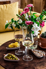 Aperitif set. Two glasses of white wine and a variety of snacks olives, pistachios, potatoes chips on a wooden table with a bouquet of flowers in a cozy room
