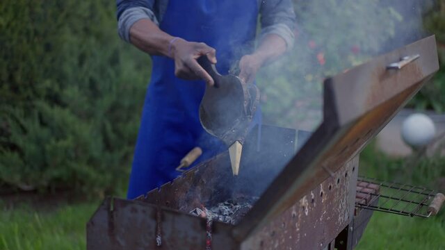 Unrecognizable African American young man in apron blowing bbq smoke outdoors on summer day. Chef using manual tool setting barbecue fire on backyard. Cooking and cuisine concept