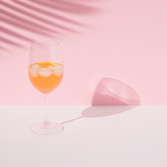 A cold orange cocktail with ice in a tall glass, its shade and the shade of a palm tree. Summer concept on white pink background