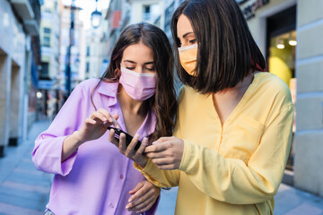 Female hispanic friends, wearing mask, search destination and shopping with smartphone. Texting and using navigator holding a mobile phone. Lifestyle portrait yellow and lavender year color