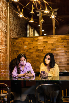 Hispanic young female friends using smartphone technology in restaurant to order dinner and chatting. Wearing face mask. Lifestyle from independent women together. Yellow and lavender color 2021