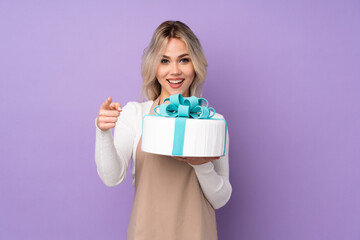 Young pastry chef holding a big cake over isolated purple background points finger at you