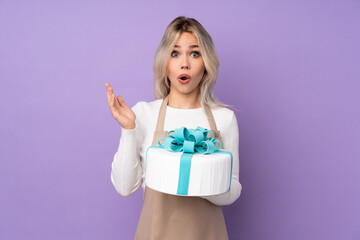 Young pastry chef holding a big cake over isolated purple background with surprise facial expression