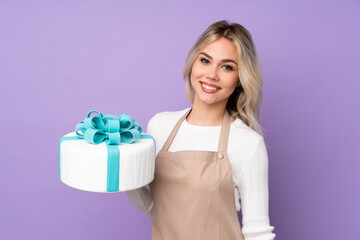 Young pastry chef holding a big cake over isolated purple background