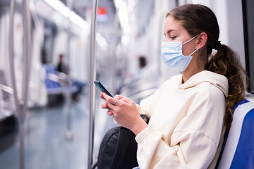 Young stylish woman in a protective face mask with mobile phone traveling by train on subway
