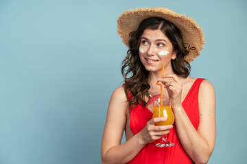 On a blue background, an attractive girl in a straw hat holds orange juice. Beautiful girl looks to the side at the copy space