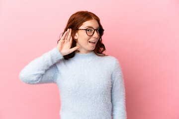 Teenager reddish woman isolated on pink background listening to something by putting hand on the ear