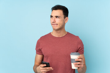 Young caucasian handsome man isolated on blue background holding coffee to take away and a mobile while thinking something