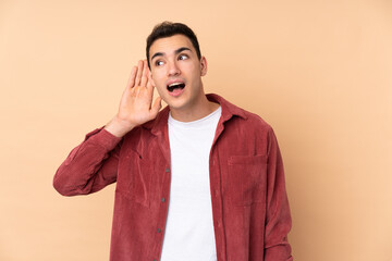 Young caucasian handsome man isolated on beige background listening to something by putting hand on the ear