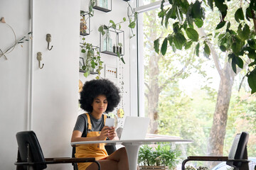 Young happy gen z black student girl with afro hair sitting at table in cafe indoor alone using mobile shop marketing app online with computer working typing surfing internet. - obrazy, fototapety, plakaty