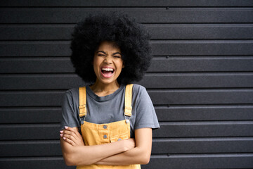 Portrait of young black afro American laughing joyful model in her 20s. Funny smiling generation z mixed race teen lady with arms crossed and eyes closed standing on black wall background.