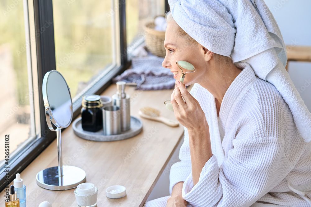 Poster gorgeous mid aged woman of 50s massaging face skin sitting in bathroom near window using natural fac