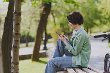 SIde view young woman 20s in green jacket jeans sit on bench near bicycle in city spring park outdoors using hold mobile cell phone chat online in social network People active urban youthful concept