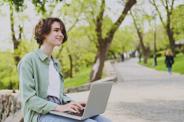 Young side view happy student freelancer woman in green jacket jeans sit on bench in spring park...