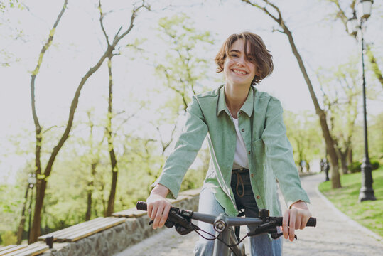 Young sporty smiling happy woman 20s wear casual green jacket jeans riding bicycle bike on sidewalk in city spring park outdoors, look camera. People active urban healthy lifestyle cycling concept