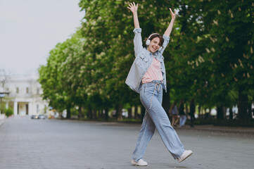 Fototapeta na wymiar Full length young fun happy student excited caucasian woman 20s wear jeans clothes headphones listen to music walking strolling down green park alley outdoors dancing People urban lifestyle concept.