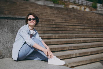 Side view young student fun woman in jeans clothes headphones eyeglasses listen to music leaning on building wall sit on concrete steps outdoors walk look camera rest. People urban lifestyle concept.