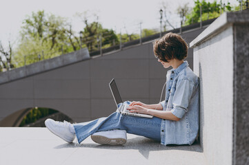 Side view full length young freelancer woman in jeans clothes headphones glasses listen to music...