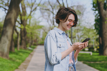 Side view young smiling caucasian fun woman 20s in jeans clothes stand in city spring park outdoors use mobile cell phone chat with boyfriend on social network People active urban lifestyle concept