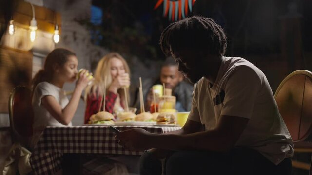 Side view of smiling African American young man texting on smartphone as blurred family talking laughing eating picnic dinner at background. Happy relaxed people enjoying barbecue outdoors
