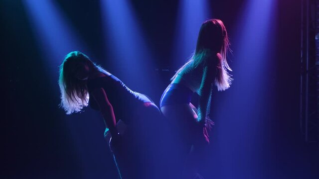 Two young hot women sexually move their bodies and twerk their ass. Silhouette of dancers in short shorts. Twerk duet on the background of blue lights. Slow motion.