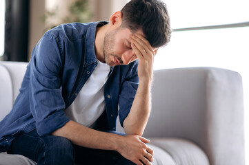 Upset sick stressed young caucasian man in casual clothes sits on sofa in the living room, bending over, massaging his head, suffering from pain, migraine, closing his eyes, needs treatment and rest