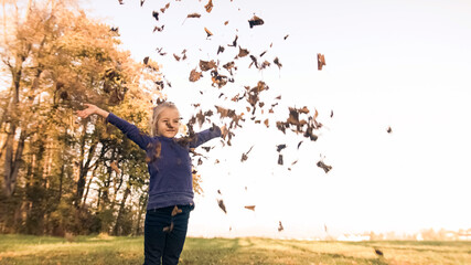 Wide shot of a smiling preschool girl, standing at the edge of the forest, holding a pile of dry leaves in hands, throwing it in the air