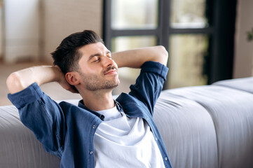 Close-up calm happy handsome caucasian bearded man in casual clothes relaxing on sofa at home, resting in living room, dreaming of vacation, put his hands behind his head, close his eyes, smiling
