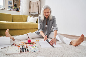 Senior female artist painting in her living room, while sitting at the floor
