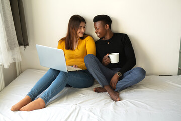 beautiful young couple african man and caucasian woman sit on bed with laptop 