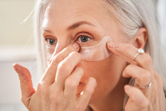 Female is looking into the mirror while wearing eye patches against wrinkles - Powered by Adobe