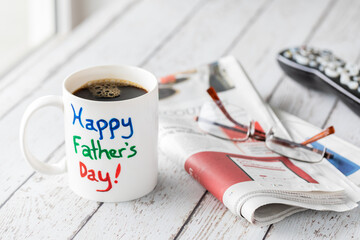Mug with Happy Father's Day painted on it filled with coffee, beside a newspaper and remote. Fathers Day concept.