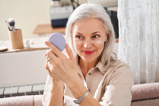 Woman with under eye patches looking at the hand mirror while painting her lips