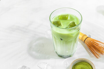 Glass of iced green matcha latte on white tile background with hard shadows of palm leaves. Cold summer drink