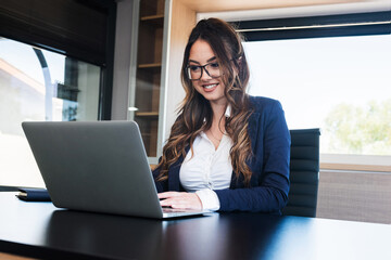 Positive business woman with brown hair working on laptop in office, wearing glasses and smiling.